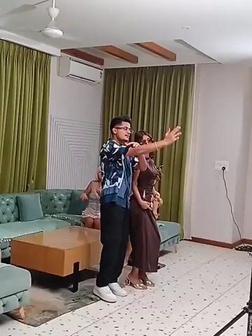 The video shows a man and a woman posing for photos in a living room setting. The man is wearing a patterned short-sleeved shirt, black pants, and white sneakers. He is gesturing with his arms. The woman is wearing a long brown dress and gold high heels. She has sunglasses on and is posing with her hands on her hips and head. Several women are sitting on the blue sectional sofa in the background. There is a coffee table in front of the sofa. The room has green curtains, a white ceiling with a ceiling fan, and a white wall with a geometric design.  Professional lighting equipment and a photographer are also visible in the room. The camera pans to show the photographer and lighting equipment, then back to the man and woman posing.