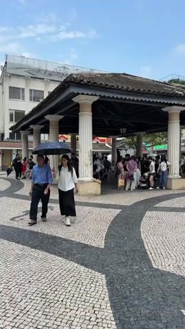 The video shows a wide angle shot of a cobblestone square in Macau, with a covered pavilion in the center. The pavilion has a dark roof supported by white columns and is filled with people, many of whom are standing. Some are sitting on the edges of the pavilion, appearing to rest or wait. Outside the pavilion, a few people are walking in different directions, including a man and a woman walking side by side. The woman holds an open umbrella. The sky is blue with a few clouds, and the buildings in the background are varied in color and style, including yellow, white, and brown. Some signs written in English can be seen on the buildings. The ground is paved with a patterned cobblestone design with darker stones creating curved pathways through the square.