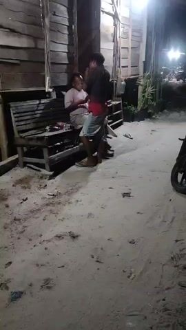 The video shows two people outdoors at night. One person, wearing a pink shirt and shorts, is sitting on a wooden bench. The other person, wearing a brown shirt and denim shorts, is standing and appears to be dancing or exercising. The environment looks like a sandy area next to a wooden building. There is a motorcycle parked on the right side of the frame. Streetlights are visible in the background.
