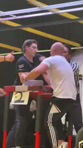 The video shows two men engaged in an arm-wrestling match on a table. The man on the left is wearing a black t-shirt and glasses, while the man on the right is wearing a white t-shirt and black Adidas pants. A referee in a striped shirt stands nearby, observing the match closely. The arm-wrestling table is red and black, and there's a number "2" sign on it. The background consists of a black curtain, a white ceiling with yellow beams, and a few onlookers. One person is giving a thumbs-up.