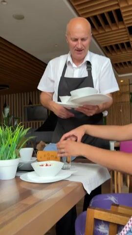 A waiter, a middle-aged bald man wearing a white shirt and a black apron, is serving food at a table in a restaurant. He is holding a stack of plates and bowls. In front of him, on the table, are two bowls of food, possibly a breakfast dish like yogurt with fruit and granola, or porridge. A woman's hands are reaching for one of the bowls. The table is set with a white tablecloth, silverware, and glasses of water, one with a lemon slice. A potted plant with green leaves is also on the table, next to a cup and saucer. In the background, the restaurant's interior is visible, featuring wooden slatted walls and ceiling, and a dark hood over a fireplace or stove. The lighting suggests it is daytime, and the atmosphere appears relaxed. A small sign on the table reads "AROMA 97 CAFFE & RESTAURANT". The waiter's expression is one of concentration as he serves the food. The woman's hands suggest she is about to begin eating.