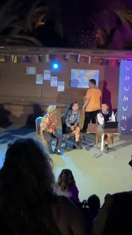 A group of four people are on a stage in front of a wooden wall adorned with flags and maps. The person furthest to the right is a woman wearing a black and white jacket, sitting at a table with a laptop and papers. To her left is a young woman in a black and white patterned top, sitting on a chair. Next to her is a man in a colorful shirt and hat, also seated. The person furthest to the left is a young man in an orange t-shirt, standing and gesturing towards the map on the wall. The audience is visible in the foreground, with some individuals in silhouette. The lighting is dim, with stage lights illuminating the performers and a blue light casting a glow on the map. Palm trees are visible in the background, suggesting an outdoor or semi-outdoor venue. The overall scene suggests a performance, possibly a play, skit, or presentation, taking place at night.