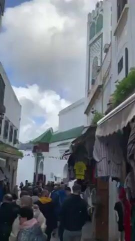 The video shows a street scene filled with people. The street is lined with white buildings, some with green tiled roofs. A tall white tower with green accents stands prominently in the background. Small shops line one side of the street, displaying clothing and other items for sale. There are flags of different nations hung between the buildings. Many people are walking along the street.
