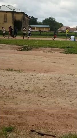 A group of individuals, appearing to be young men or boys, are engaged in a running activity. They are dressed in athletic wear, including shorts and t-shirts, in various colors like red, yellow, blue, and white. The scene is outdoors, on what appears to be a dirt track or path, with a dusty, unpaved surface.

In the background, there are buildings, including a light yellow, two-story structure with visible windows and a damaged roof. Other buildings with darker roofs and what seem to be residential houses are also present. Vegetation, including trees and grass, borders the running area and the buildings. The sky is overcast, suggesting a cloudy day, and the time of day appears to be daytime, likely afternoon, given the light.

The individuals are spread out along the track, with some running in a line and one person sitting on the ground to the far right. Their postures suggest exertion and movement, indicating they are either training, participating in a race, or involved in some form of athletic exercise. The overall atmosphere seems to be one of physical activity within a somewhat rural or underdeveloped setting. No text is visible in the media file.
