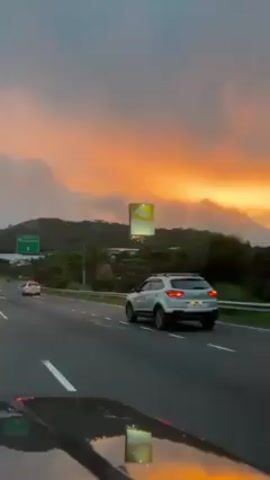 Here is a description of the video:

The video captures a scene from the driver's seat of a car traveling on a highway as the sun sets. The sky is filled with vibrant oranges and pinks, diffused by patches of clouds, creating a dramatic backdrop. Road signs indicate directions to "La Zona," "Salida," and "Colon" and a large billboard is visible along the roadside.

The car is moving along the multi-lane highway, with other vehicles visible in adjacent lanes and ahead. The road is marked with white lines, and there are streetlights illuminating the highway. The horizon features a stretch of green, hilly terrain.