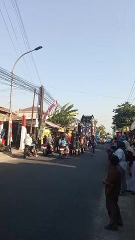 A street is filled with people and motorbikes. A large decorated truck with lights and sound equipment is moving down the street, followed by a crowd of people and motorbikes. People are standing on the side of the street watching the procession. The sky is clear and blue. The buildings on either side of the street appear to be shops or houses. Flags and banners are visible, suggesting a festive event.
