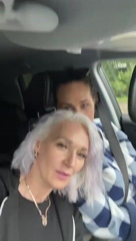 Two women are in the front seats of a car. The woman in the foreground has short, light purple hair and is wearing a black top and a necklace. She is looking at the camera and smiling, making a hand gesture. The woman in the background has dark hair and is wearing a blue and white checkered shirt. She is looking at the camera and making a peace sign with her fingers. The car appears to be moving, as indicated by the motion blur. The environment outside the car is green and appears to be a rural or wooded area.