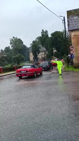 A person in a bright yellow rain suit directs traffic on a wet asphalt road. Three cars are visible: a red convertible in the foreground facing away, a dark car behind it, and a white hatchback to the left. In the background, several buildings and lush green trees line the road, with a stone bridge to the left. The sky appears overcast.