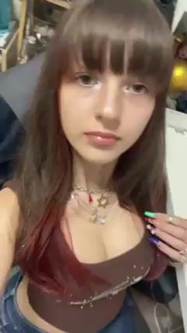 A young woman is recording a selfie video. She has long brown hair with bangs and is wearing a brown tank top and blue jeans. She is also wearing several necklaces and rings. She points to the rings on her fingers and then gestures her hand in the air. The background is cluttered with various items, including a cabinet and a black leather garment. Music is playing in the background.