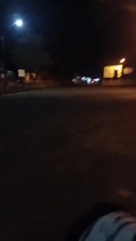The video is taken at night from the perspective of someone riding a motorcycle. A street lamp illuminates a paved area in front of a building with lights on. The motorcycle's dashboard is visible, showing a lit display. The rider is wearing a jacket with a white and black patterned design. The scene is dark with some distant lights and structures.