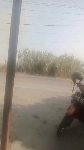 The video shows a person riding a motorcycle on a dusty road. The motorcycle is black and the rider is wearing a white helmet and a gray shirt. The road is unpaved and there is a lot of dust in the air. In the background, there are tall grasses and some power lines. Another motorcycle is parked in the foreground on the right side of the frame. The sky is hazy and the lighting suggests it is daytime.