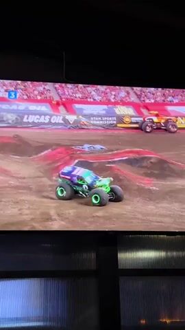 The video shows a screen displaying a monster truck rally. A green and purple monster truck with the name "Grave Digger" is airborne, mid-jump over a dirt obstacle. Other monster trucks are visible in the background, lined up. The screen displays a timer that reads "00:00:00" and "00:00:01". Below the screen, a dark corrugated surface with horizontal lines of light is visible. The overall scene suggests a broadcast of a live event.