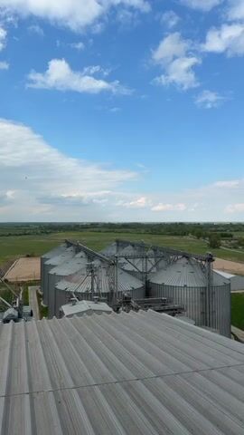 The camera pans slowly across the top of a group of metal grain silos and the surrounding farmland under a partly cloudy, blue sky. The silos are connected by metal conveyor systems. Beyond the silos, a flat landscape of green fields and scattered trees extends to the horizon. A gray, rectangular building with several windows stands on a green lawn next to the silos. A road runs through the landscape, and cars can be seen driving on it. The foreground of the video shows the corrugated metal roof of a structure from which the video is being taken.
