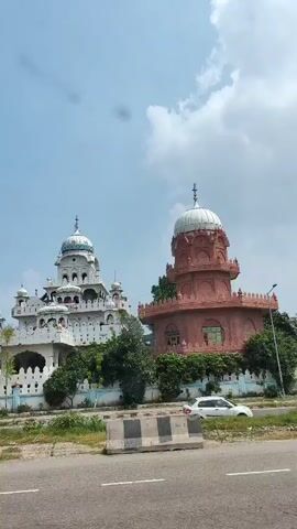 The video shows a white car driving on a road. On the left side of the road is a large white building with multiple domes and intricate white decorative elements. To the right of the white building is a smaller, reddish-brown building with a dome. The sky is cloudy. There are some trees and vegetation visible between the road and the buildings. The scene appears to be in a sunny, outdoor environment.