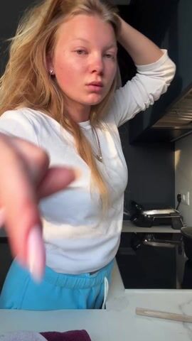 A young woman is standing in a kitchen. She has long blonde hair, light skin, and is wearing a white long-sleeved shirt and light blue sweatpants. She is posing, touching her hair and placing a hand on her hip. In the background is a kitchen counter with a cooktop, an electrical outlet, and a range hood. There is also a grill and a pot on the counter. Sunlight is shining into the kitchen.