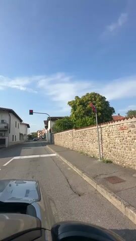 The video begins from the perspective of someone in a vehicle, looking forward down a street. The vehicle is stopped at a red traffic light. To the left are the sides of buildings, and to the right is a stone wall with a tree behind it. The sky is blue with some clouds. The video continues for a short duration, and then the traffic light turns green. The vehicle begins to move forward.
