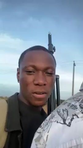 The video shows two Black men standing outdoors. The man in the foreground, wearing a dark jacket over a black shirt, looks directly at the camera. Behind him and slightly to his right, another man wearing a white shirt and a black baseball cap with a red bill is partially visible. A black pole, possibly a light stand, is positioned behind the first man. The sky is visible in the background, with a mix of blue and white clouds. In the distant background, there are trees and what appears to be a road or path. The audio suggests a windy environment.