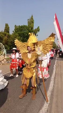 The video shows a parade on a sunny day. In the foreground, a person in a detailed golden eagle costume with large wings and a feathered headpiece walks down a paved road. They are holding a wooden staff. To the left and behind the eagle costume, a person in a white hat and a red and white dress is walking. Further back, several people in white and red outfits with headdresses are marching in procession. A large Indonesian flag, red and white, is visible on the right side of the frame, held by someone off-camera. Trees line the sides of the road, and the sky is clear and blue. The overall atmosphere appears festive.