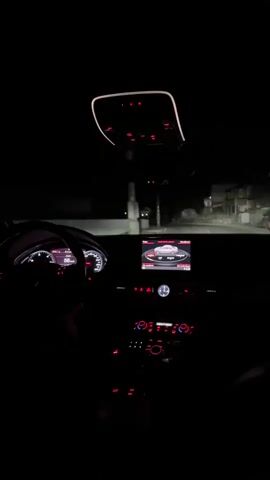 A person is driving an Audi at night. The dashboard and controls are illuminated in red. The navigation screen shows the car in a bird’s eye view. The driver’s hands are on the steering wheel. The road ahead is visible through the windshield, lit by the car’s headlights. There are buildings visible in the distance.