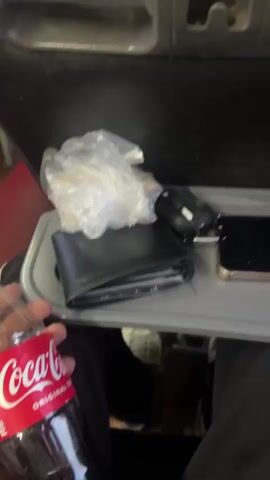The video captures scenes from inside a passenger bus, likely in Thatta Haji, Pakistan, during the daytime.

The initial seconds feature a close-up, blurred shot of a hand holding a small plastic bottle of Coca-Cola with a red cap. The background is indistinct but shows what appears to be an interior compartment of a vehicle, containing items like a white plastic bag, a dark wallet or pouch, and car keys.

The view then transitions to the interior of the bus. It features rows of dark grey seats with red accents. Overhead luggage racks run along the ceiling, with some bags and items placed on them. Red curtains are drawn over the windows. Several passengers are visible; most are seated, including one person in a light blue shalwar kameez in the foreground. A man dressed in a light grey shalwar kameez is standing in the aisle, bending slightly, possibly interacting with another passenger or adjusting something. At the front of the bus, a screen is visible, displaying the text "YUTONG," which is a known bus manufacturer, and a number "0315-7776." The bus interior is brightly lit, indicating it is daytime, and the overall setting suggests the bus is in motion.