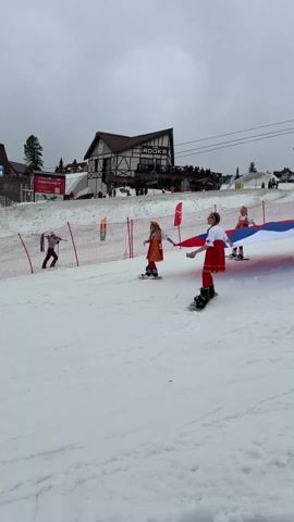 On a snowy slope with a building labeled "ROOKS" in the background, a group of individuals dressed in red and white traditional clothing, and wearing skis or a snowboard, hold up a large Russian flag. The sky is overcast. There are other people visible in the background near the building and near a safety fence to the left. Some people yell in the video, seemingly in support.