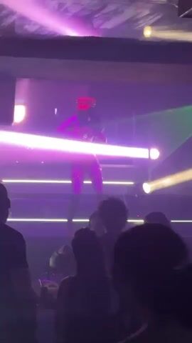 Here is a description of the video:
A performer is dancing on a stage in a nightclub, while a crowd watches. The dancer is wearing a red, sparkly outfit with a matching hat and thigh-high boots. The stage is lit with colorful lights, including blue, pink, green, and yellow, and there are also several white spotlights. There is a screen behind the performer that displays blue geometric shapes. The crowd is silhouetted in the foreground. A security guard is also visible in the crowd. The music is loud and energetic.