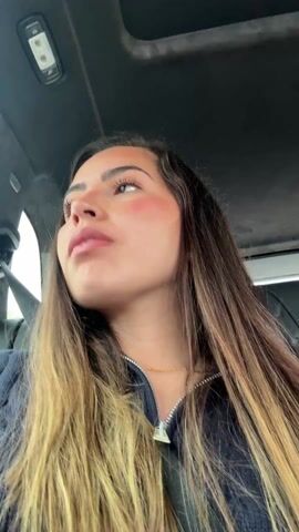 The video shows a young woman with long brown and blonde hair, wearing a dark blue jacket and a gold necklace, sitting in a car. She is looking upwards and then to the side. The interior of the car is visible, including the ceiling, a sunroof, a seatbelt, and leather seats.