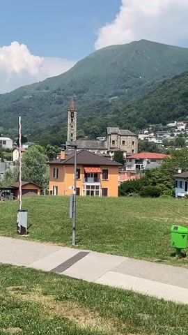 This is a video clip showcasing a scenic view of the village of Tesserete, Switzerland. The foreground features a paved walkway bordered by lush green grass. To the left, a white and red striped barrier gate stands vertically, indicating a potential entrance or exit. A light pole is positioned in the middle of the grassy area.

In the midground, an orange house with a dark roof and red striped awnings is prominently visible. Behind it, a stone church with a tall bell tower rises above the other buildings. The background is dominated by a majestic, tree-covered mountain under a bright blue sky with scattered white clouds. The village itself is nestled on the mountainside, with houses cascading down the slopes.

The overall impression is that of a peaceful, sunny day in a picturesque European mountain village. The time of day appears to be midday or early afternoon, given the bright sunlight and distinct shadows. The weather seems pleasant and warm. There is no visible text in the clip. The scene evokes a sense of tranquility and natural beauty.