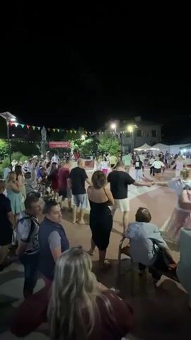 This is a vertical, wide shot video of an outdoor nighttime gathering. The scene is lit by string lights and streetlights, with a dark sky above. A crowd of people, estimated to be between 50 to 100 individuals, are participating in a circle dance. The dancers are holding hands and moving in a circular formation. Some spectators are seated and observing the dance. In the background, there are buildings and trees. The overall atmosphere appears to be festive.