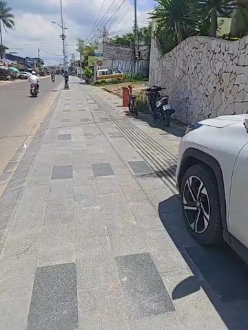 A white car is parked on the right side of the frame, partially obscuring a sidewalk. The sidewalk is made of gray square tiles with textured strips running parallel to the road. Beyond the sidewalk, a low, curved wall made of irregular white stones lines the property. Behind the wall, lush green plants, including palm trees and large-leafed tropical plants, are visible. A white, modern-looking house with an orange-tiled roof is situated behind the vegetation. On the left side of the frame, a paved road stretches into the distance, with a few motorbikes and a white van. The sky is blue with scattered white clouds. The scene appears to be a street in a tropical or subtropical location on a sunny day.