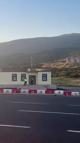 The video shows a Jandarma station in Turkey, filmed from a moving vehicle on a nearby road. The station is a single-story building with the word "JANDARMA" displayed prominently. Two people are standing outside the building: one appears to be in a Jandarma uniform, and the other is wearing a black tank top and pants. A marked Jandarma vehicle is parked nearby. In the background, a hillside is covered with apartment buildings, trees, and power lines. The sky is clear and the sun is setting.