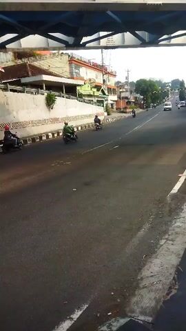 The video shows a street scene with several people riding motorcycles. The road is asphalt and appears to be a busy street with some traffic. There are buildings on the left side of the road and trees and other vegetation on the right. The sky is visible and appears to be overcast. The video is shot from a slightly elevated perspective, looking down on the street.