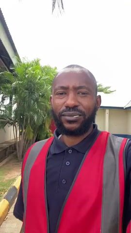 The media file shows a close-up, selfie-style video of a Black man with a beard, wearing a navy blue polo shirt and a red and gray high-visibility vest. He has a toothpick in his mouth. The background suggests an outdoor setting, possibly a work environment. There are palm trees and other lush green foliage visible, along with part of a building with light-colored walls and a blue trim. A yellow and black striped barrier is also present on the left side of the frame. The sky is overcast, indicating a cloudy day. The man's expression is neutral. The location is identified as Kinkinau, Nigeria.