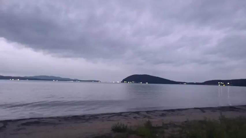 The video shows a wide view of a body of water, likely a bay or ocean, under a cloudy sky. In the foreground, there is a sandy shore with some scattered vegetation. Across the water, there are several hilly landforms, possibly islands or peninsulas, with small lights visible on them, suggesting human habitation or structures. The water is relatively calm, with gentle waves lapping at the shore. The lighting suggests it is either dawn or dusk, with the sky being overcast and the overall scene appearing muted and slightly dark. There are no people or discernible activity in the video.