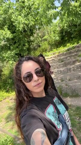 Two women are outdoors on a sunny day amidst lush green foliage and stone steps. The woman in the foreground is filming herself with a smartphone, smiling at the camera. She has long, curly dark hair and is wearing sunglasses, a black t-shirt with a graphic design, and a fanny pack. Her left arm, visible in the shot, has a tattoo. Behind her, another woman with dark, shoulder-length hair and glasses is standing on a dirt path, looking at her own smartphone. She is wearing a maroon t-shirt, black pants, and white sneakers. Stone steps are visible on a slight incline behind the second woman, suggesting a natural or historical site. The overall atmosphere is bright and pleasant, indicating good weather. The scene takes place in Vilafranca del Penedès, Spain.