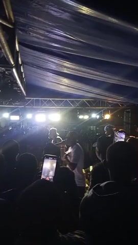 A low-angle, medium shot video taken at night shows a concert crowd viewed from behind. The foreground is filled with the heads of concertgoers. In the mid-ground, on a stage illuminated by bright white lights from above, a performer wearing a white t-shirt and a baseball cap is visible, holding a microphone and speaking or singing. To the left of the performer, another person is also on stage, wearing a dark jacket and a white hat, facing the crowd. A large, dark tarp is stretched overhead, supported by metal scaffolding. The audience is silhouetted against the stage lights, with many holding up cell phones to record the performance. The overall atmosphere suggests a lively and energetic musical event.