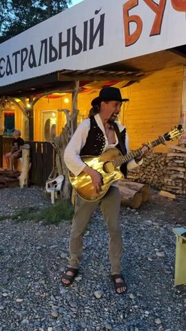 A man in a cowboy hat and vest plays an acoustic guitar and sings in front of a wooden building with a sign that reads "ТЕАТРАЛЬНЫЙ БУФЕТ" (Theater Buffet). He is wearing a white shirt, a dark vest, khaki pants, and sandals. His left hand is on the fretboard of the guitar, and his right hand is strumming. He is looking towards the camera and appears to be singing. Behind him, there are stacked logs and a person sitting on steps. The ground is covered in gravel. The lighting suggests it is dusk or evening.