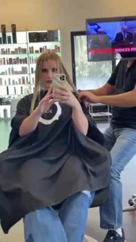 The scene is inside what appears to be a hair salon. A young woman with light blonde hair is sitting in a salon chair, wearing a black cape. She's holding a smartphone and taking a selfie. A man, presumably the hairstylist, is standing behind her, cutting her hair with scissors. He's wearing a black t-shirt, light blue jeans, and black sneakers. A television screen is visible in the background, showing what appears to be a news broadcast in a language other than English. Shelves stocked with hair products are visible along the wall behind the woman. The room is well-lit, and the floor appears to be tiled.