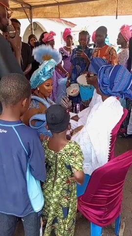 Here is a concise description of the video:

The video shows a gathering of people, seemingly at an event or celebration. Many individuals are dressed in vibrant, colorful traditional clothing. Some people are taking photos or videos with their phones, and others are interacting with the person seated, possibly offering blessings or gifts. There is a bucket or container nearby, likely for collecting contributions. The setting appears to be outdoors, under a tent-like structure. The atmosphere is likely celebratory and community-oriented.