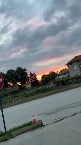 The video captures a static outdoor scene at sunset in Dolna Oryahovitsa, Bulgaria. The sky dominates the upper half of the frame, showcasing a dramatic blend of dark grey clouds and vibrant streaks of orange, pink, and yellow from the setting sun. Silhouetted against this colorful backdrop are several trees, including a prominent conifer on the left and various deciduous trees.

In the mid-ground, parts of what appear to be old or slightly dilapidated houses with tiled roofs are visible, interspersed with green vegetation and some untended ground. A light pole can be seen on the right side of the frame, further indicating a settlement. The foreground features a paved road or path with cracks, and a white and red striped barrier or tape runs horizontally across the lower part of the frame, suggesting a boundary or construction area. No people or specific activities are visible, emphasizing the natural beauty of the sunset over the landscape.