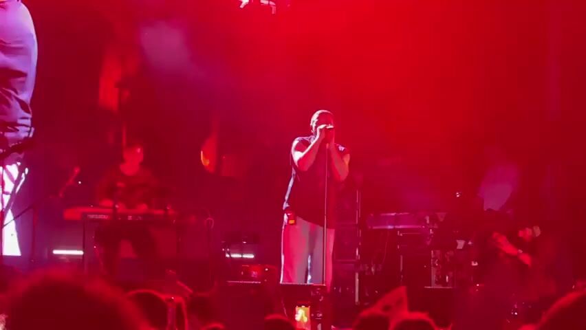 A live concert is underway, illuminated by vibrant purple and red stage lights and a haze of smoke. In the center, a male singer, wearing a sleeveless dark t-shirt and light-colored pants, holds a microphone and gestures with his right hand. To his left, a person wearing glasses and a dark shirt plays a red keyboard. To the singer's right, another person is seated, holding a camera and looking towards the stage. In the background, other band members and equipment are visible, including additional keyboards and audio equipment. The audience is visible in the foreground, with many holding up their phones to record the performance. The atmosphere is energetic, with smoke machines creating dramatic visual effects.