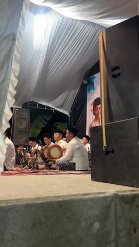 A group of young men, dressed in white robes and black skullcaps, are seated on a red and white rug on a raised stage. They are playing various hand drums, which are white with wooden rims. An older man in a white robe and patterned sarong is seated to the right of the group, also holding a drum. In the background, there are large black speakers and a white tent structure with a chandelier-like light fixture hanging from the ceiling. To the far right, a poster is visible featuring a young boy in a similar outfit. The lighting is dim, with the primary light source coming from the chandelier and some ambient light from the poster. The overall scene appears to be an indoor performance or gathering.