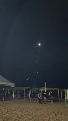 The video shows a nighttime beach scene with fireworks. In the foreground, there is a sandy beach with several people sitting on chairs and benches. There are also some tents or canopies set up on the beach. In the background, fireworks are exploding in the dark sky. One large firework is in the center of the frame, exploding into a bright, starburst pattern. Other smaller fireworks are visible in the sky as well. A red glow from a firework is visible near the ground.