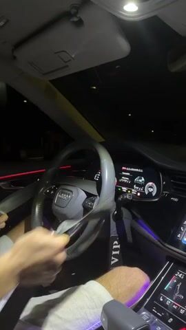 A person is shown inside a car at night, wearing light-colored shorts. Their hands are on a grey steering wheel, and they are fastening a grey seatbelt. The car's interior is illuminated by red and purple ambient lighting, with multiple digital screens visible on the dashboard displaying car information and a navigation map. A dashcam is mounted on the windshield, and a "VIP" tag hangs near the rearview mirror. The car then begins to drive, with the view through the windshield showing blurry streetlights and a dark road, indicating movement at night.