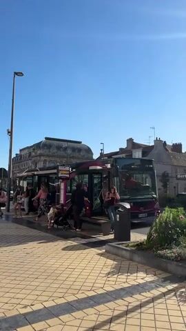 The video shows a sunny day with a clear blue sky. Two buses, predominantly maroon and black with pink accents, are parked at a bus stop. Several people are visible, some waiting to board the buses, others already on them. One bus has its doors open, and a few individuals are disembarking. Another bus also appears to have passengers inside. People of various ages are present, including adults and children, some with strollers. The bus stop area is paved with beige and gray checkered tiles. A tall lamppost stands to the left of the frame, and behind the buses, a large, ornate building with a classical architectural style is visible. The scene appears to be in an urban or town setting, with other buildings and street elements in the background. The overall atmosphere seems calm and ordinary for a public transportation hub on a bright day.