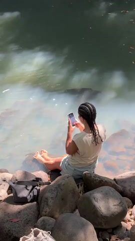 A person with braided hair sits on rocks by a body of water, holding a smartphone. They are wearing a light-colored t-shirt and denim shorts. A black bag is on the rocks to their left. The water is murky with white foam and reflects sunlight and shadows. The background shows a rocky cliff face with a waterfall cascading down.