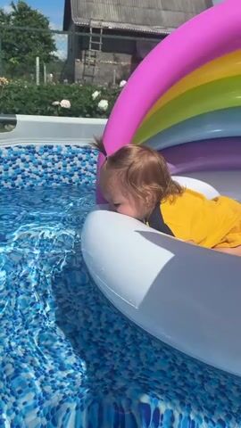 A toddler with light brown hair pulled into a small ponytail, wearing a yellow shirt with dark trim, lies on an inflatable pool float shaped like a rainbow. The toddler is leaning forward, with their face close to the edge of the float, seemingly interacting with the water. The float is in a swimming pool with a blue and white patterned liner. The background shows a wooden fence, greenery, and a building under a bright blue sky. The sun is shining, casting shadows.