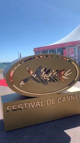 The video shows a large sign for the Festival de Cannes in France. The sign is gold in color and features the festival's iconic palm frond logo. It is placed on a block also featuring the text "Festival de Cannes". In the background, a white tent labeled "Boutique Officielle" is visible, along with a glimpse of the Mediterranean Sea under a clear blue sky. People can be seen walking and standing in the background, near a red carpet. A bird flies across the top of the screen.
