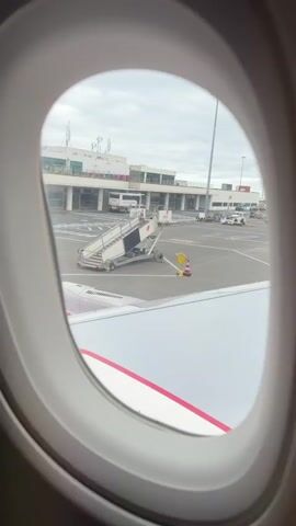 From the perspective of an airplane window, the video shows an airport terminal and tarmac. The airport building is visible in the background with a white facade, glass windows, and multiple entry points. Several pieces of ground service equipment are on the tarmac, including two aircraft stairs, one clearly labeled "portway handling de portugal s.a." and marked with the number "23-09". The plane's pink engine cowling and a red and white arrow painted on the wing are also visible in the foreground. The sky is overcast. There are people visible at the terminal. The plane appears to be taxiing, as the ground service equipment becomes larger in frame.