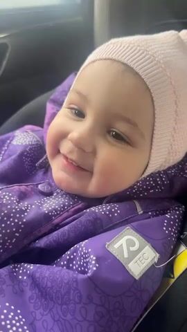 A young child with dark eyes and rosy cheeks is seen up close, smiling and laughing softly. The child is wearing a light pink knitted hat and a vibrant purple winter suit or overall, which features white polka dots and a subtle patterned design. A white label on the left arm of the suit is visible, displaying "R TEC". The child appears to be seated in a car seat or similar carrier, with a dark background suggesting an indoor or vehicle setting. The activity is the child expressing joy and amusement through their laughter and wide smile, revealing small teeth. The overall mood is one of happiness and warmth. The location context provided is Sadovod-Sad-3, Russia, though this is not visually discernible from the video itself.
